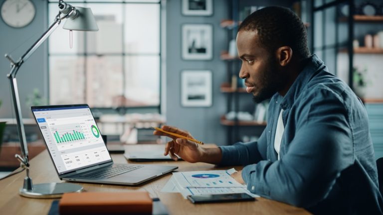 Handsome Black African American Specialist Working on Laptop Computer in Creative Home Living Room. Freelance Male is Doing Market Analysis and Creates Report with Charts for Clients and Employer.